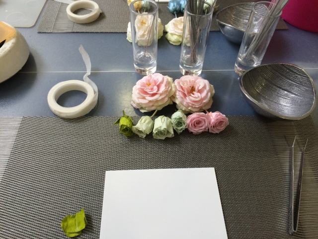 テーブルの上に並べられたピンクと白の造花、ガラス花瓶、テープや道具が置かれたフラワーアレンジメントの作業風景
